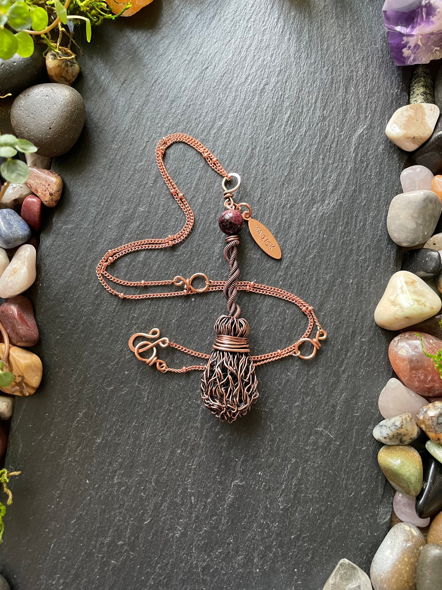 Copper necklace with a magick name tag on a dark stone surface surrounded by pebbles and small plants