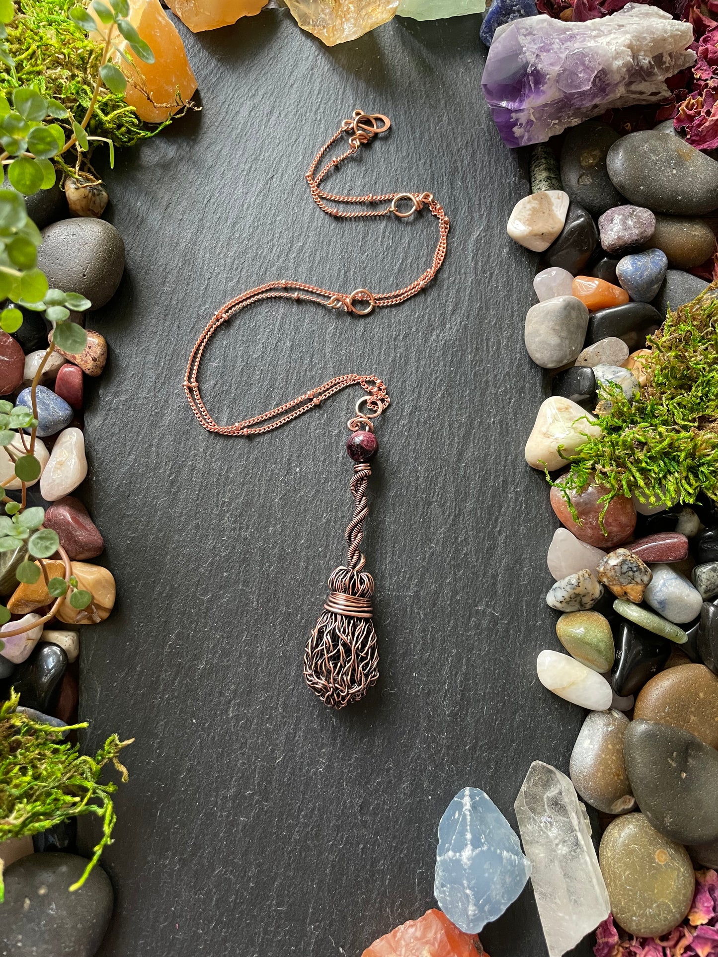 Copper pendant with decorative elements on a dark surface surrounded by stones and plants