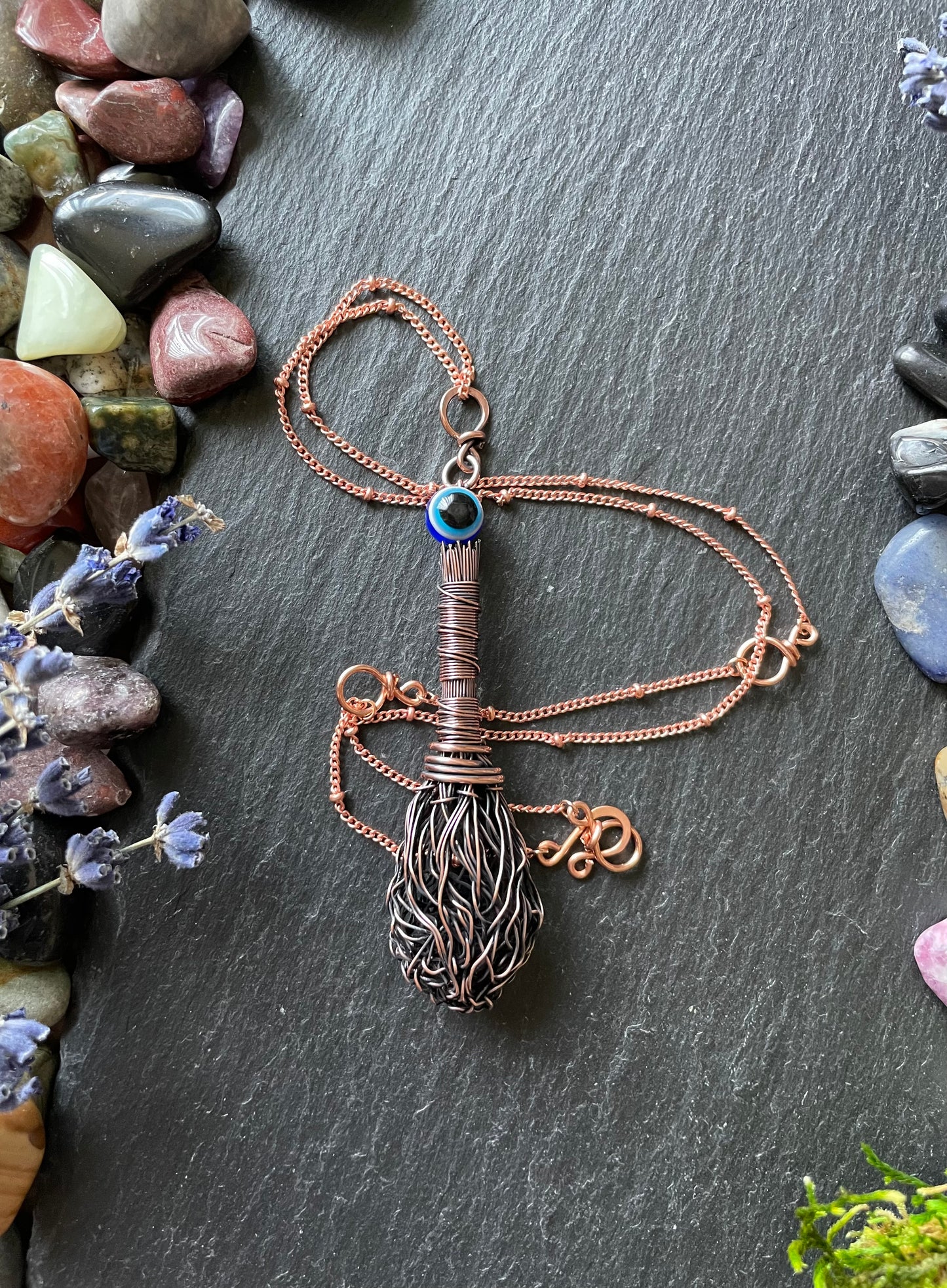 Miniature antiqued copper wire witch broom pendant with resin evil eye bead, attached to a matching copper chain and displayed on a stone slate surrounded by gemstones
