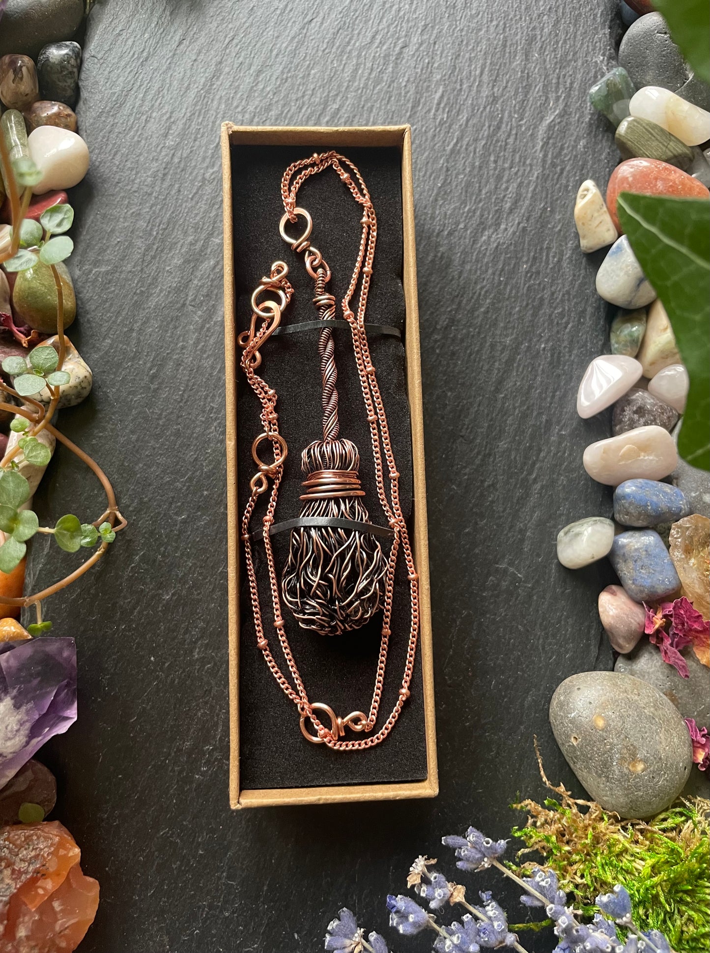 Copper broom necklace in a box on a dark surface with stones and plants around