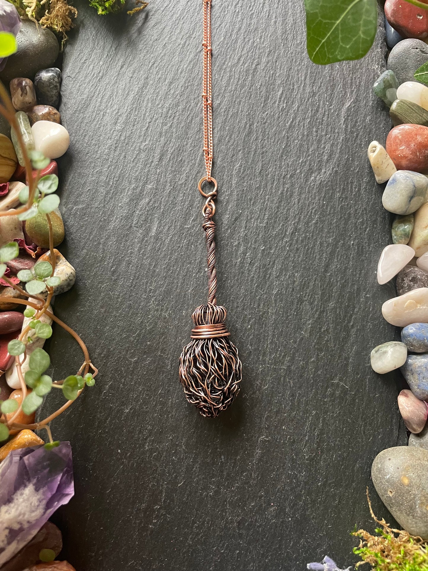 Decorative broom pendant on a dark surface with pebbles and plants.