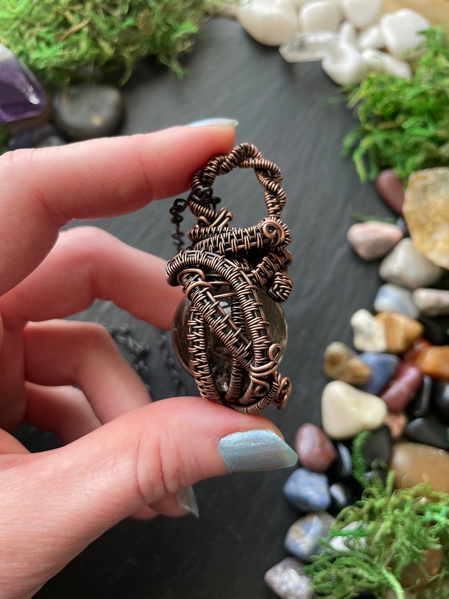 Hand holding a detailed copper wire-wrapped crystal against a natural stone and moss background