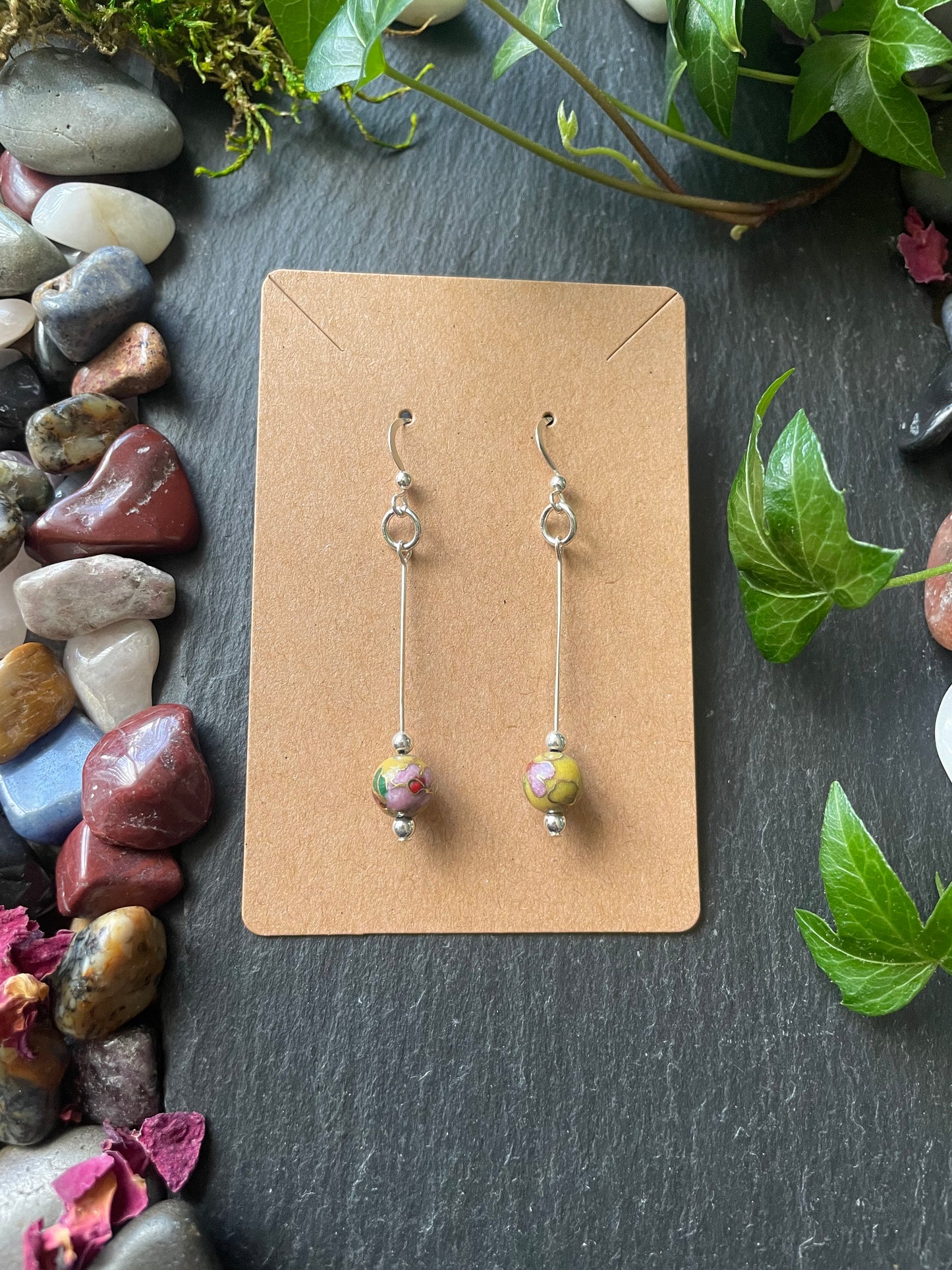 Pair of earrings on a cardboard display card with green leaves and stones in the background