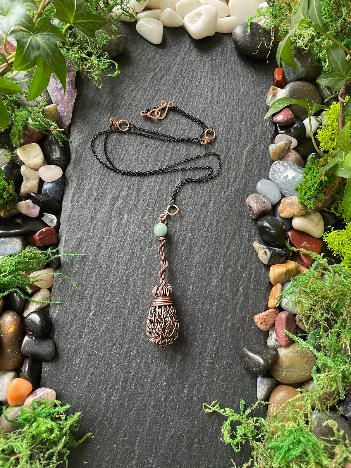 Copper broom necklace with a pendant on a slate surface surrounded by stones and greenery