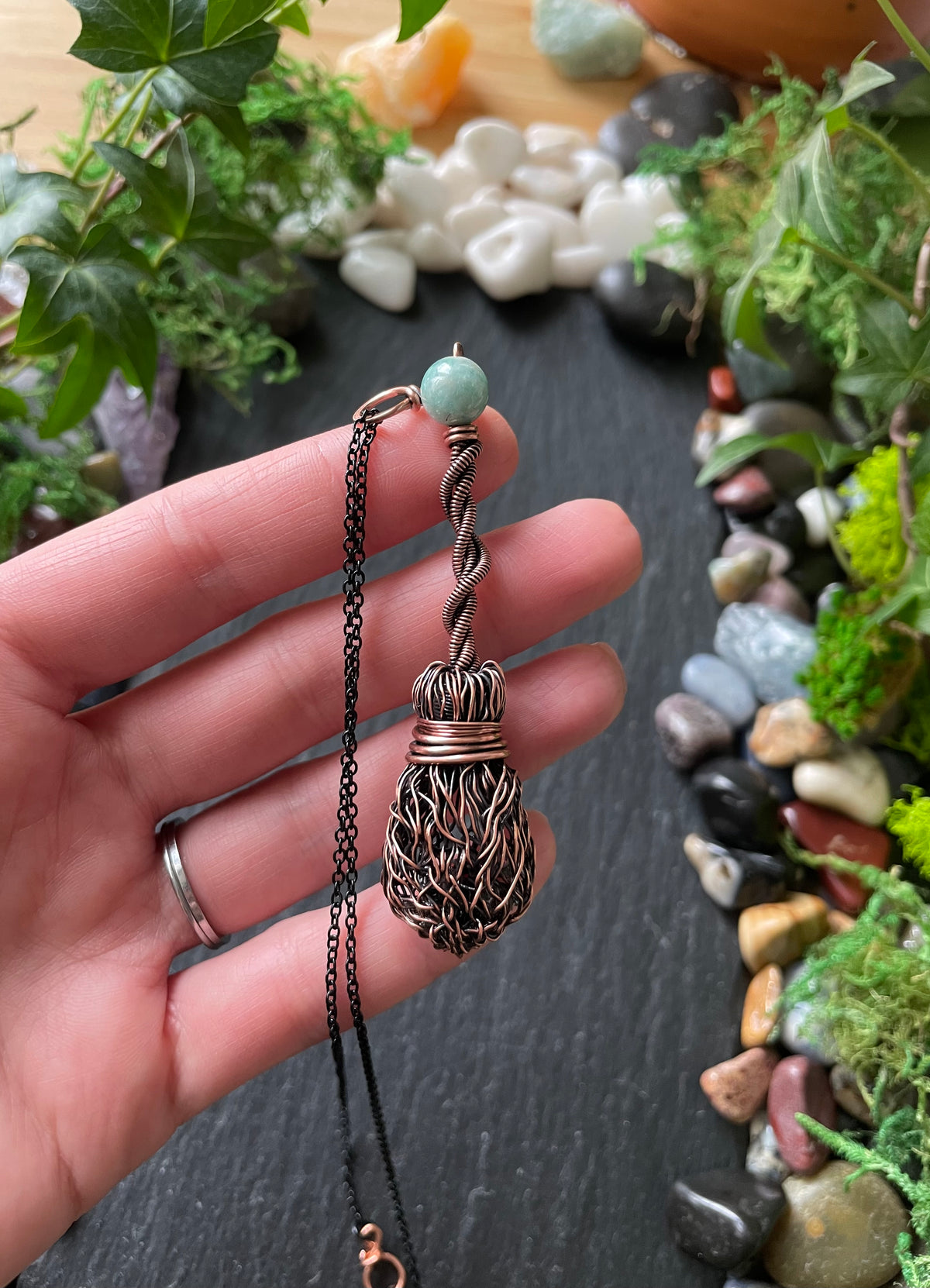 Hand holding a decorative broom necklace against a natural background of stones and plants.