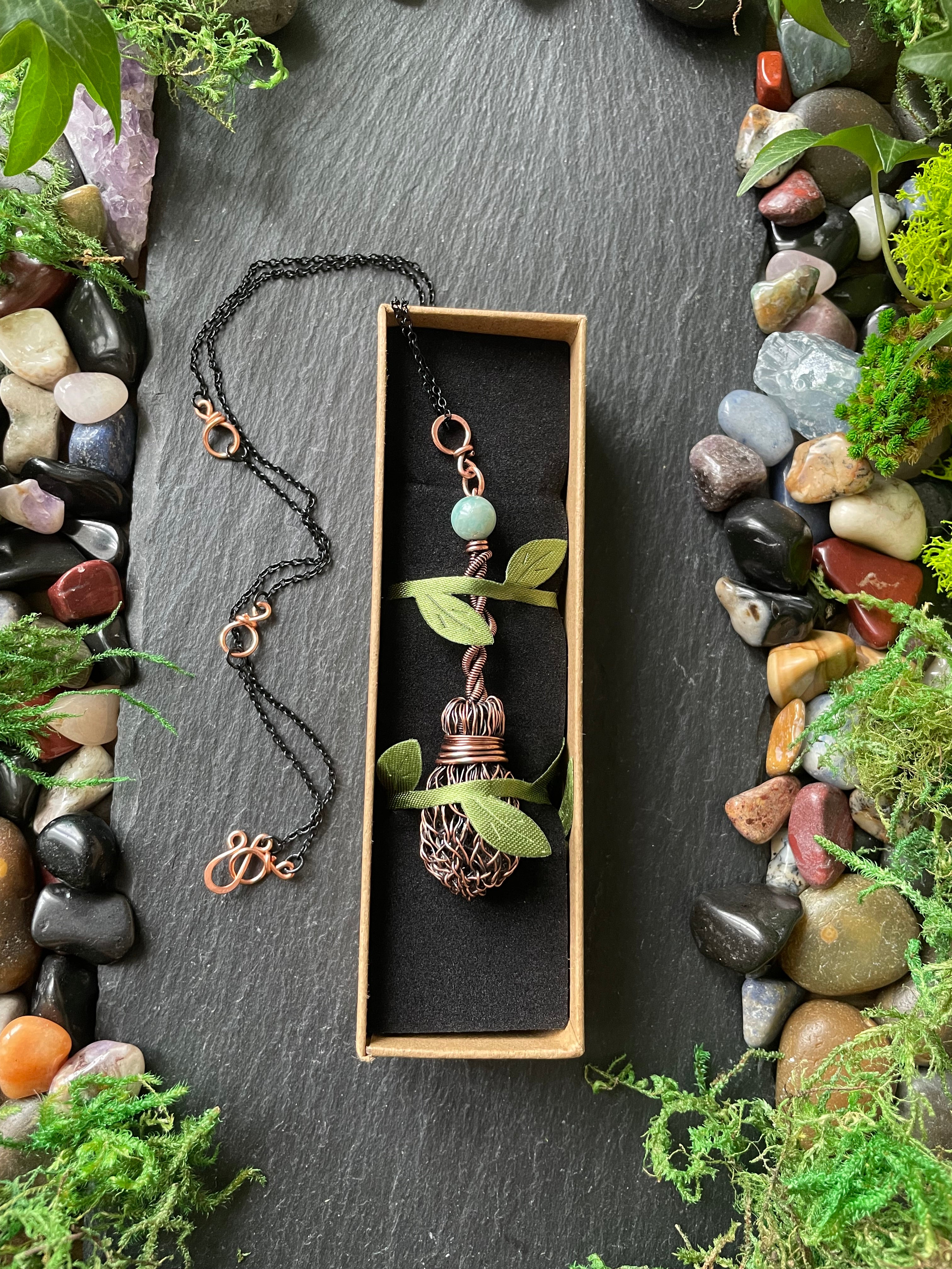 Small broom necklace with blue bead in a box on a dark surface with stones and plants.