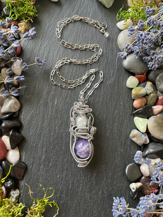 A wire wrapped crystal ball amulet with amethyst sphere and glass vial filled with clear quartz crystal, placed on a black stone slate and surrounded by colorful gemstones