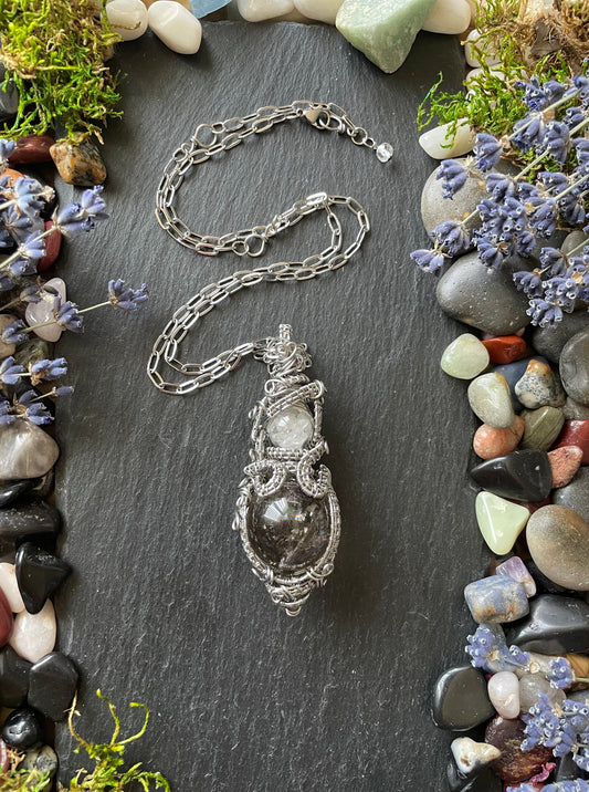 A magic crystal ball amulet with large smoky quartz sphere and glass vial filled with clear quartz crystals, in a stainless steel wire weave, and placed on a stone slate surrounded by colorful gemstones