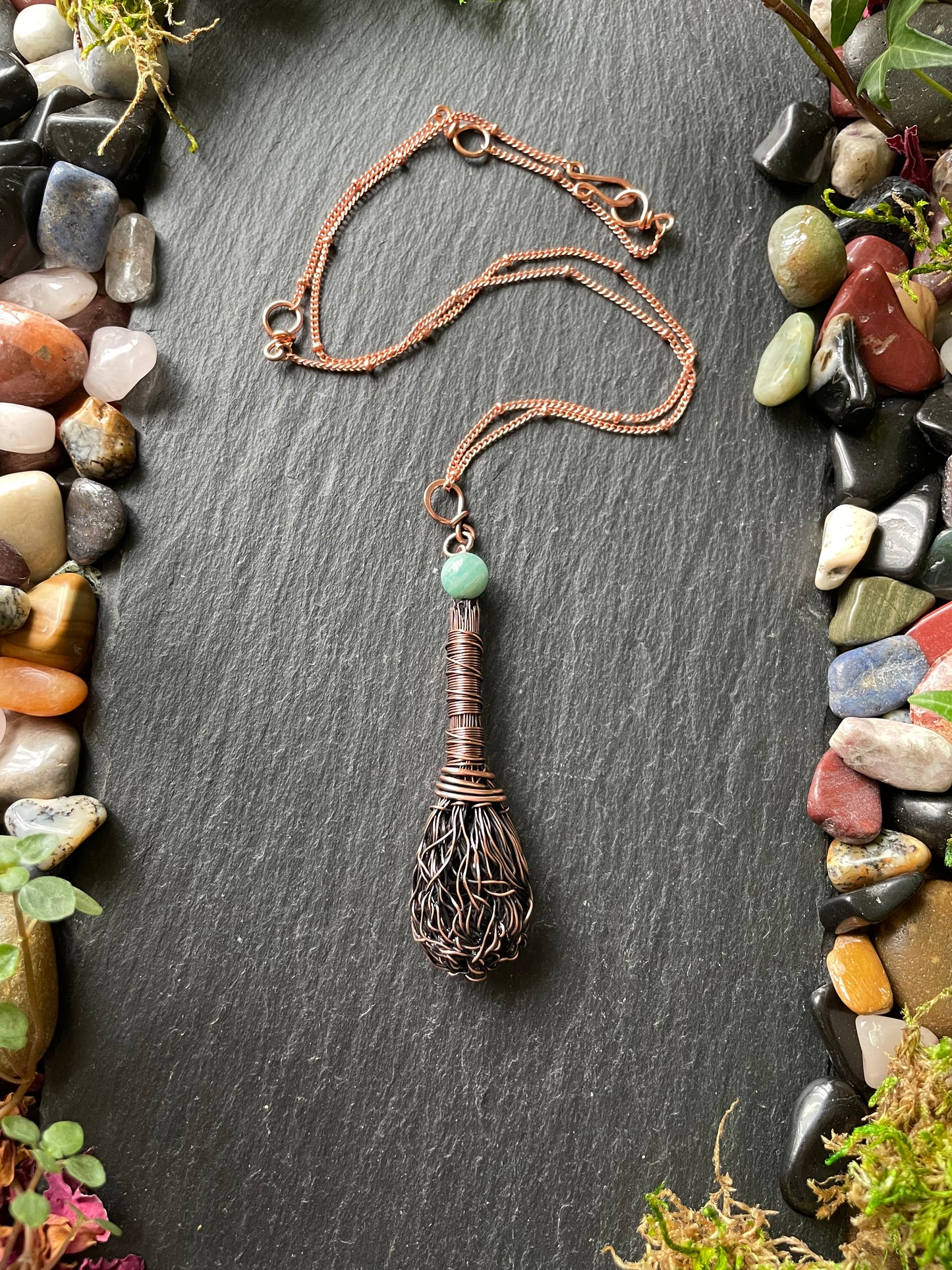 Necklace with a unique pendant on a dark slate surface surrounded by small stones and plants.