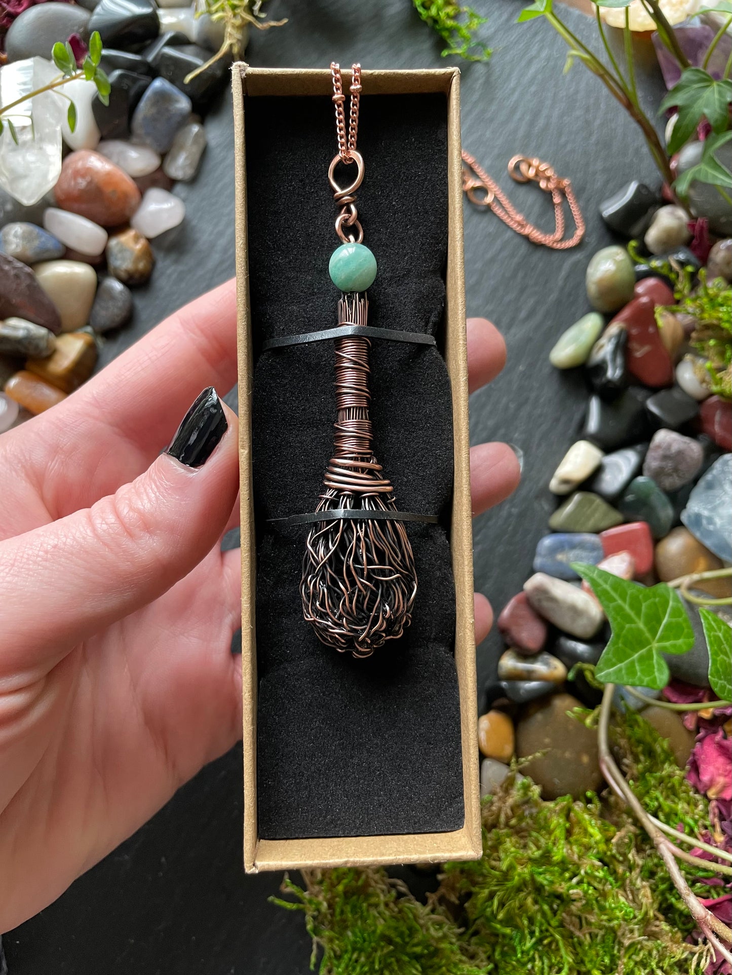 Copper wire-wrapped pendant with a green gemstone in a box, surrounded by stones and plants.