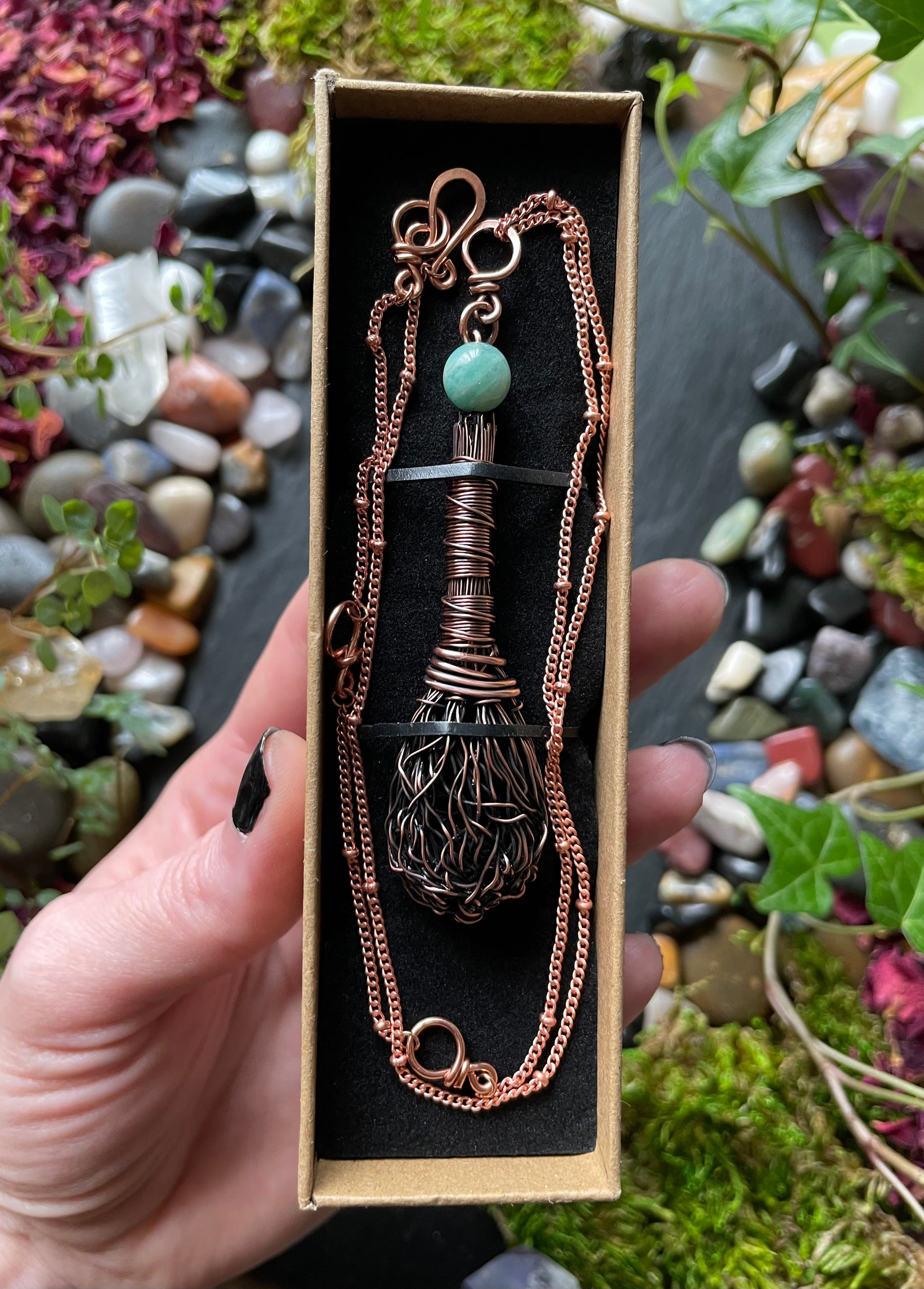 Copper necklace with a green gemstone in a box, held against a natural stone and plant background.
