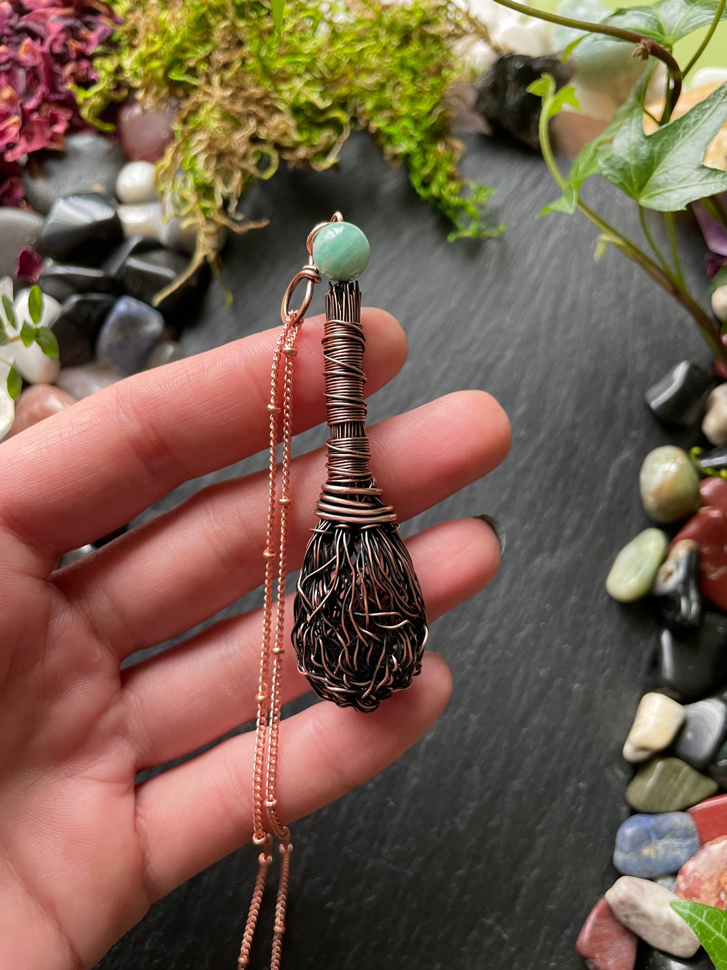 Hand holding a copper wire-wrapped pendant with a green bead against a natural stone background