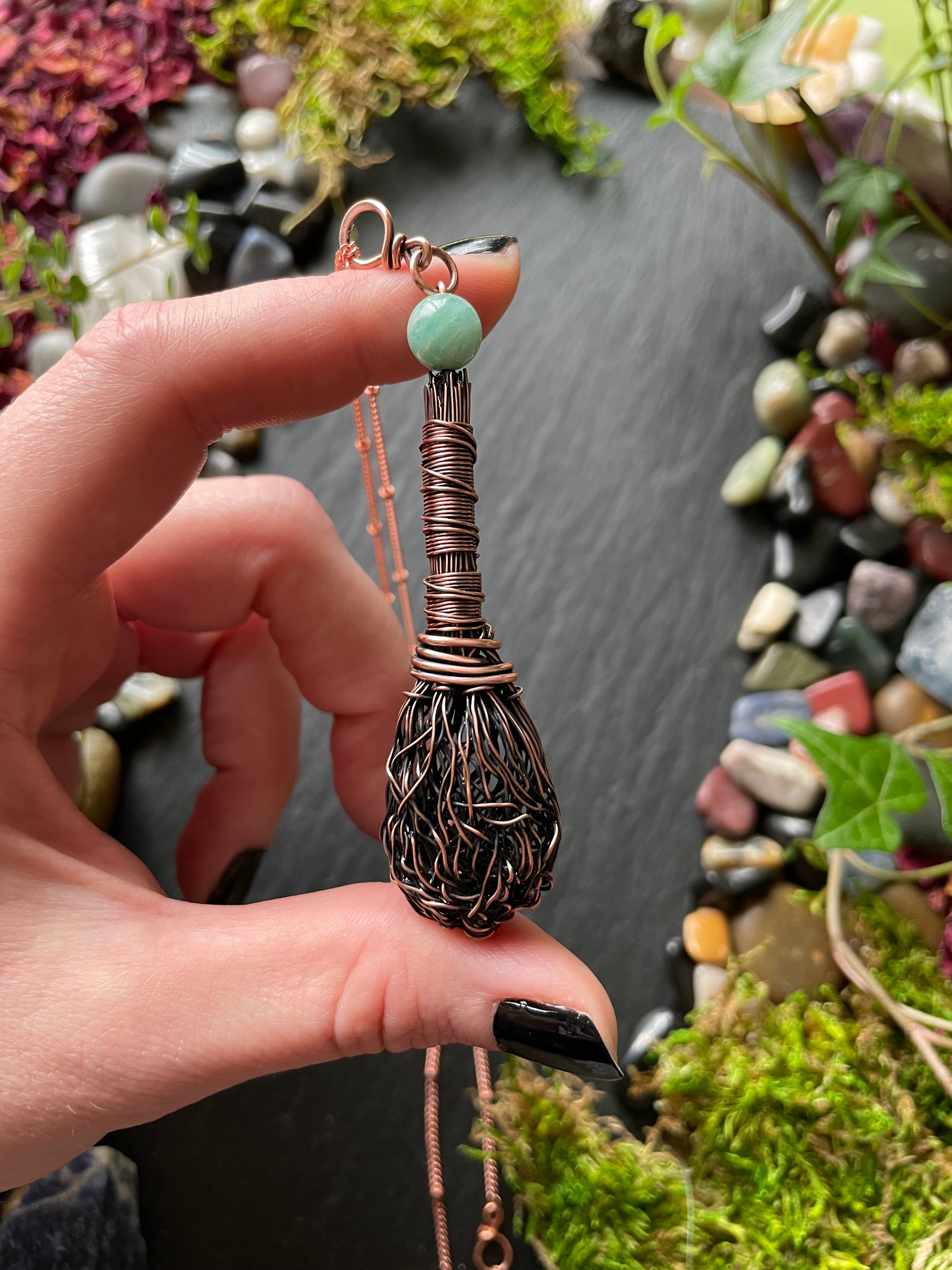 Hand holding a decorative broom with a natural stone and wire design, surrounded by stones and plants.