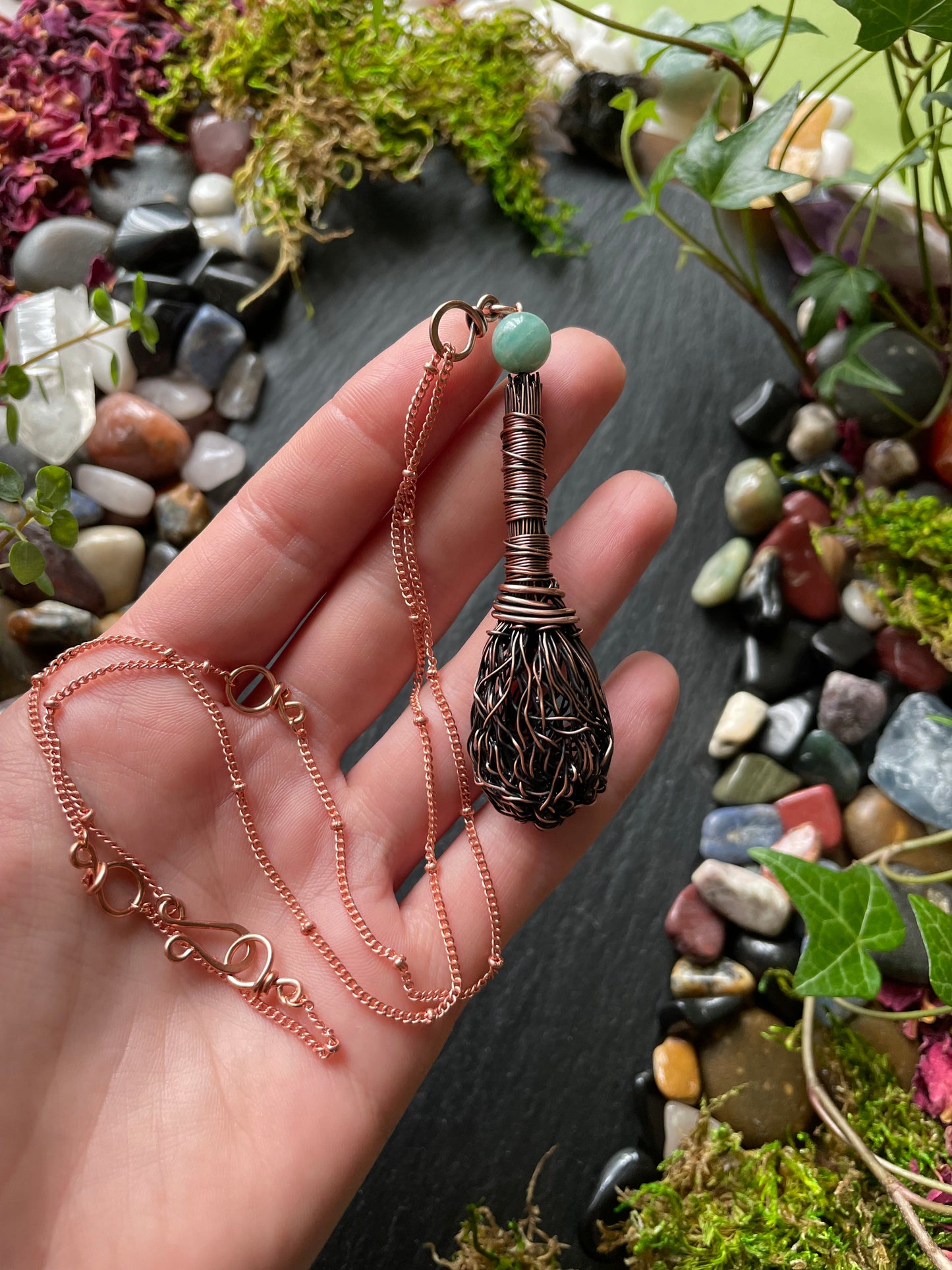 Hand holding a copper necklace with a decorative pendant against a natural background of stones and plants.