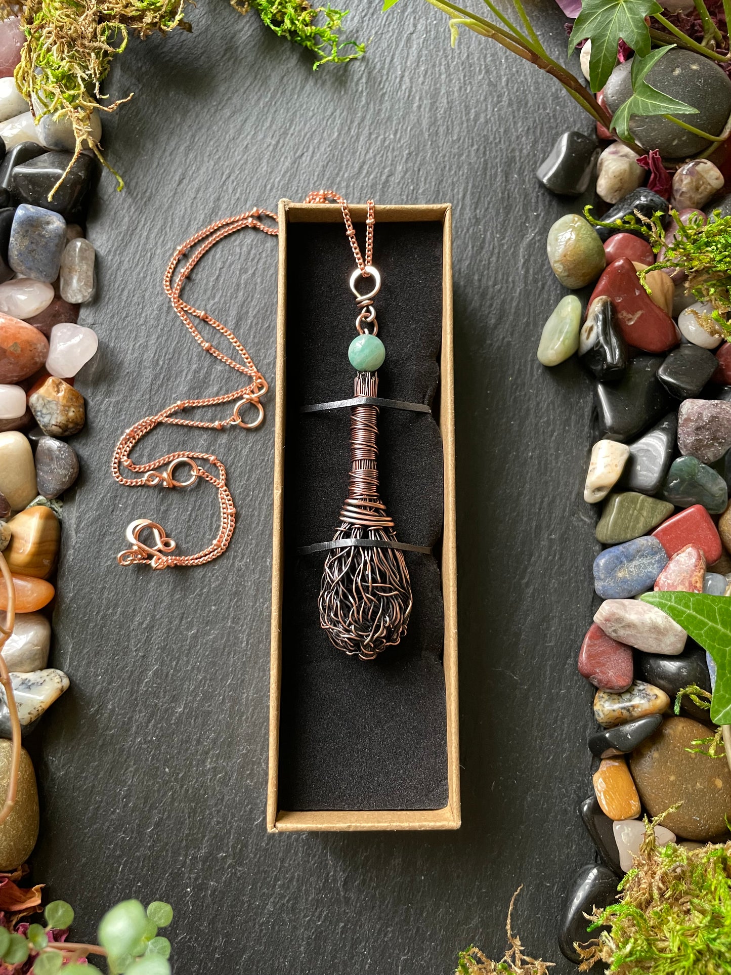 Necklace with a pendant in a box on a dark surface with pebbles and plants.