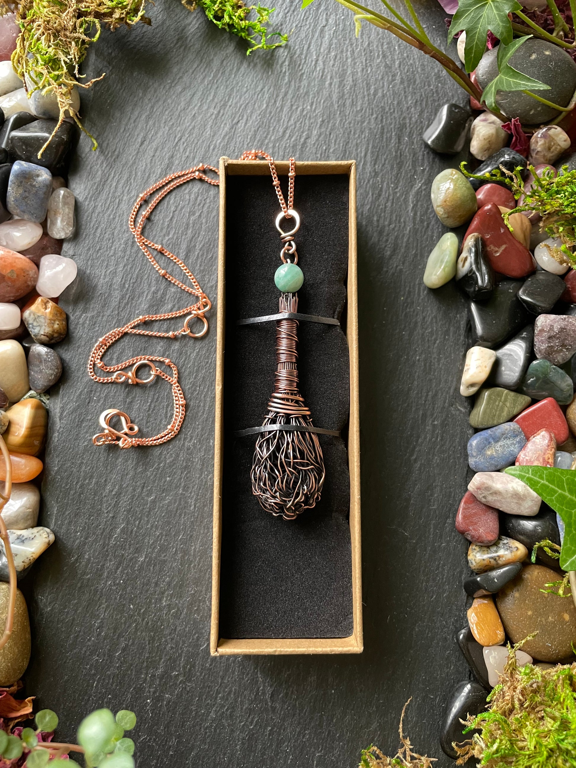 Necklace with a pendant in a box on a dark surface with pebbles and plants.