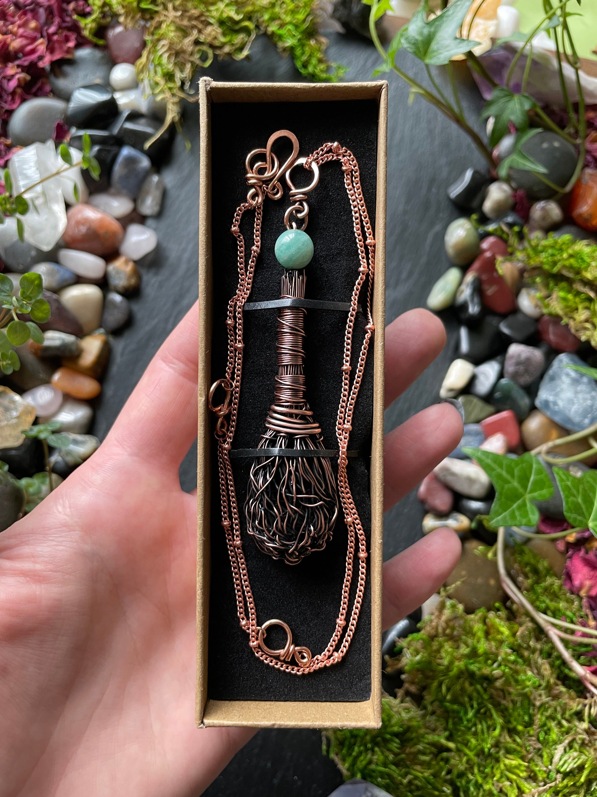 Hand holding a decorative necklace in a box with a natural background of stones and plants.