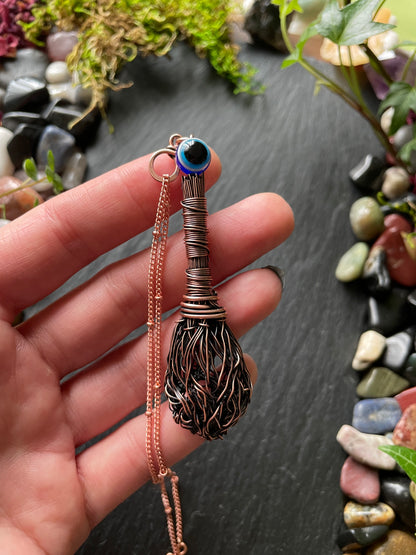 Hand holding a decorative broom-shaped pendant with a stone-like center on a dark surface with stones and plants.