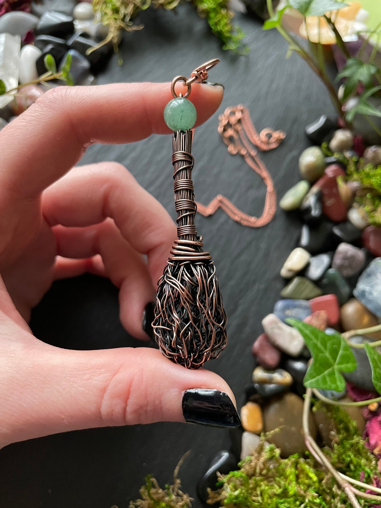 Hand holding a decorative broom with a green stone against a natural background of stones and plants.