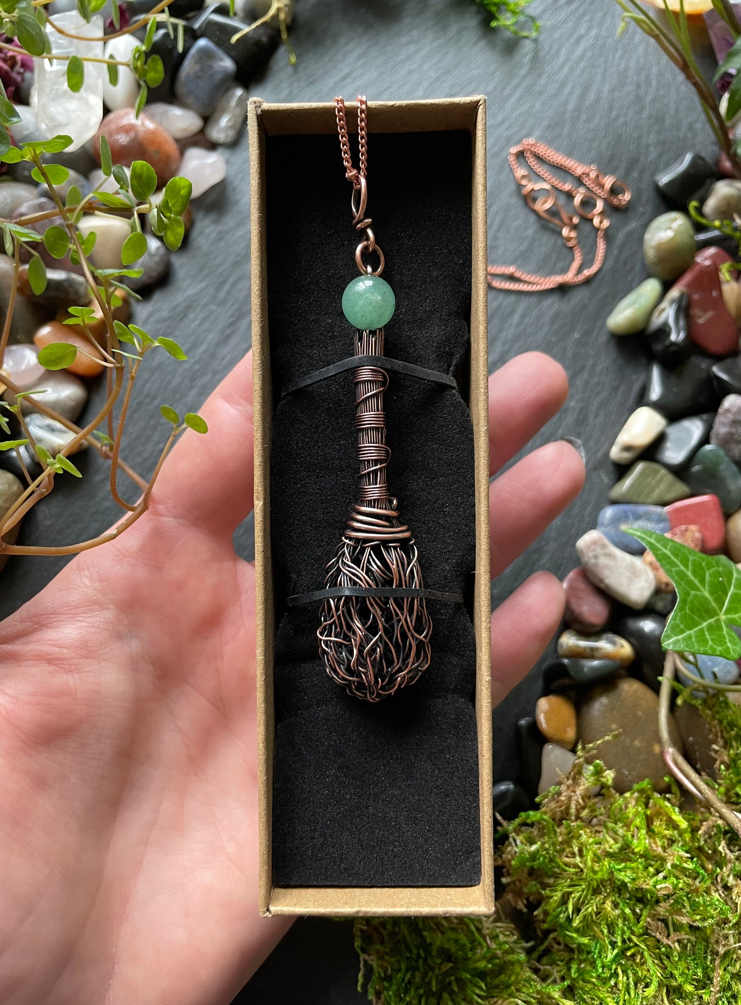 Hand holding a box with a copper and green bead necklace against a natural stone and plant background