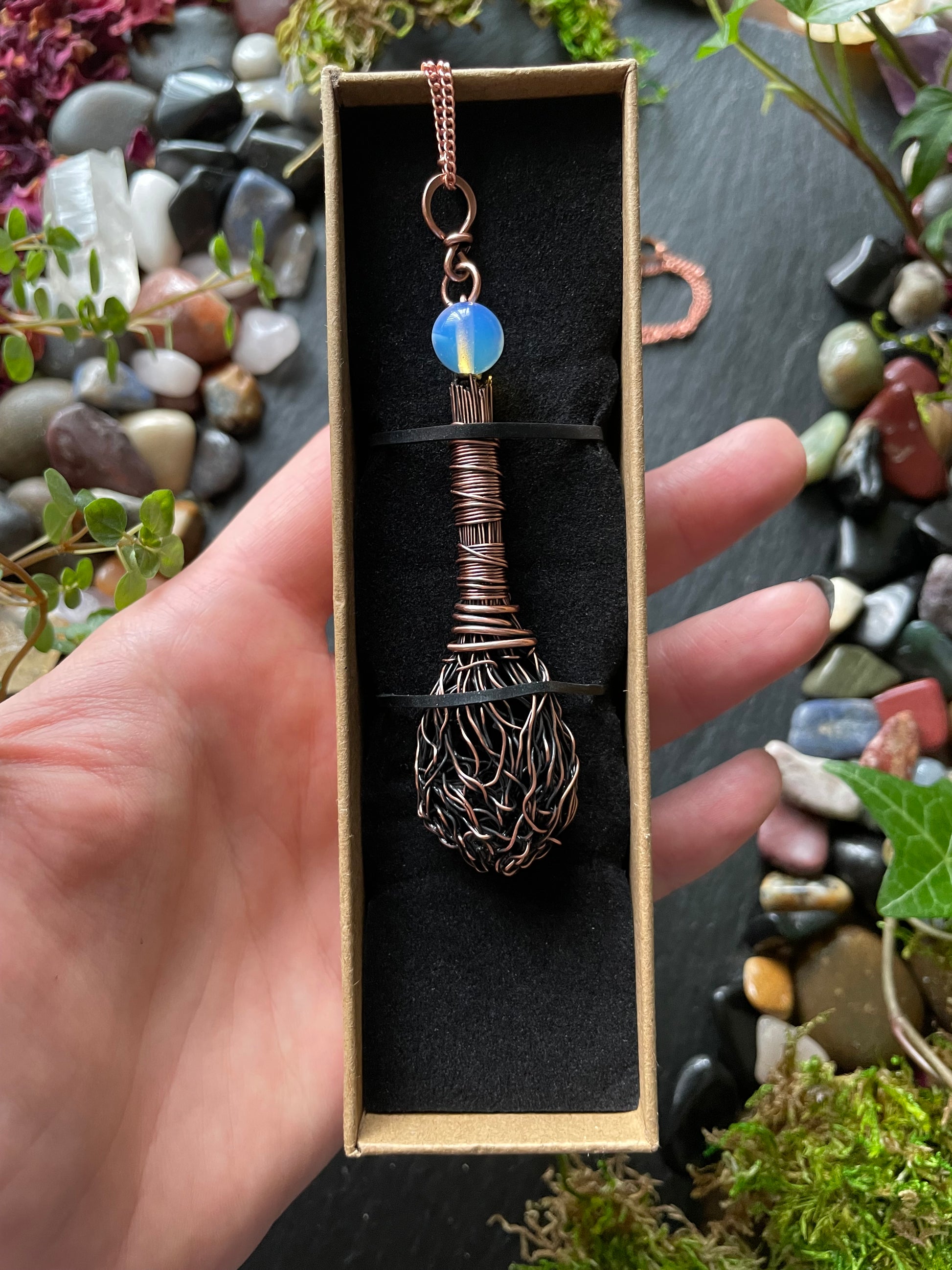 Copper broom-shaped pendant with opalite gemstone in a box held by a hand against a natural stone background.