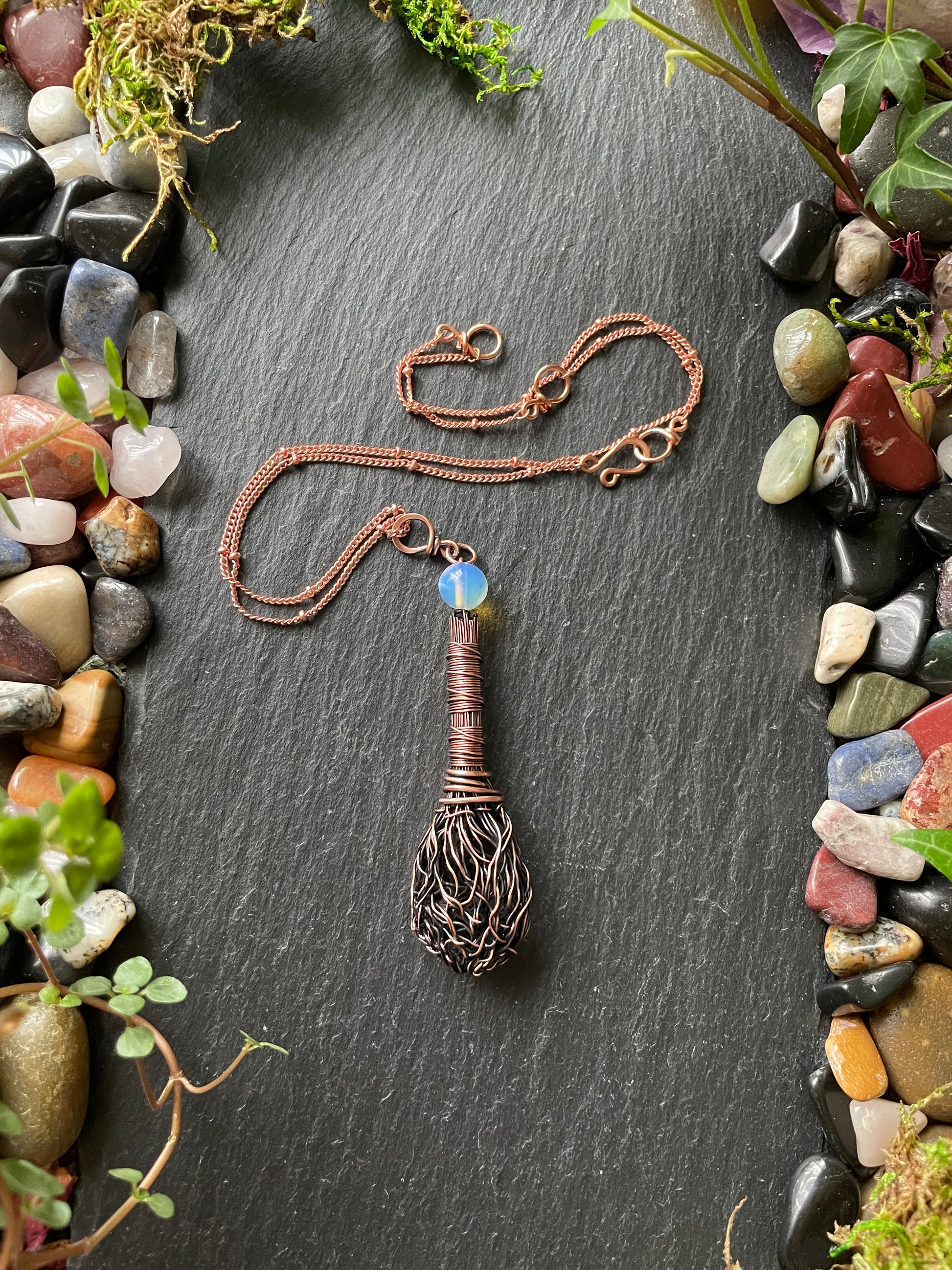 Copper necklace with a decorative pendant on a dark stone surface with pebbles and plants.