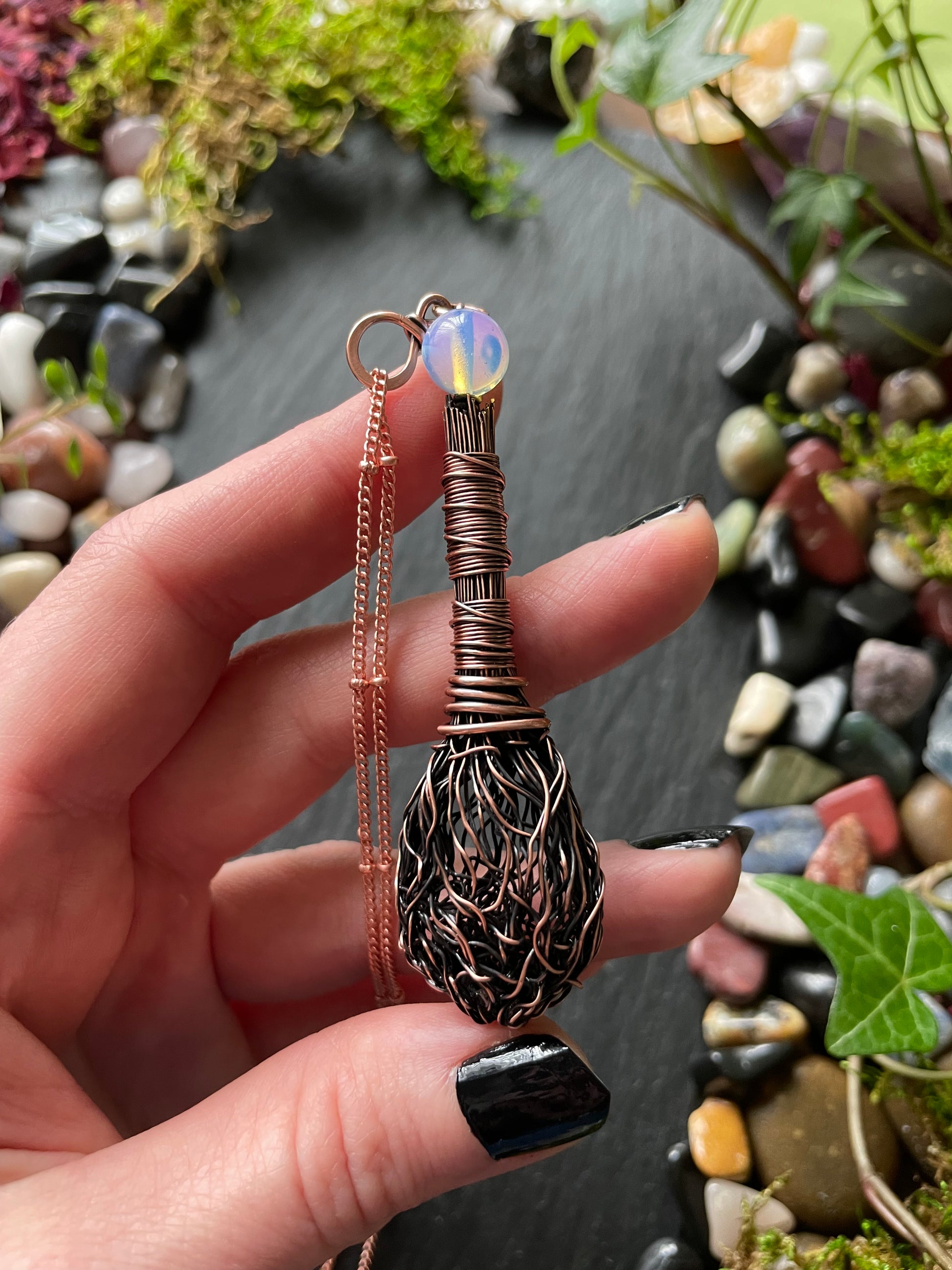 Hand holding a decorative broom-shaped object with a gemstone against a natural stone and plant background