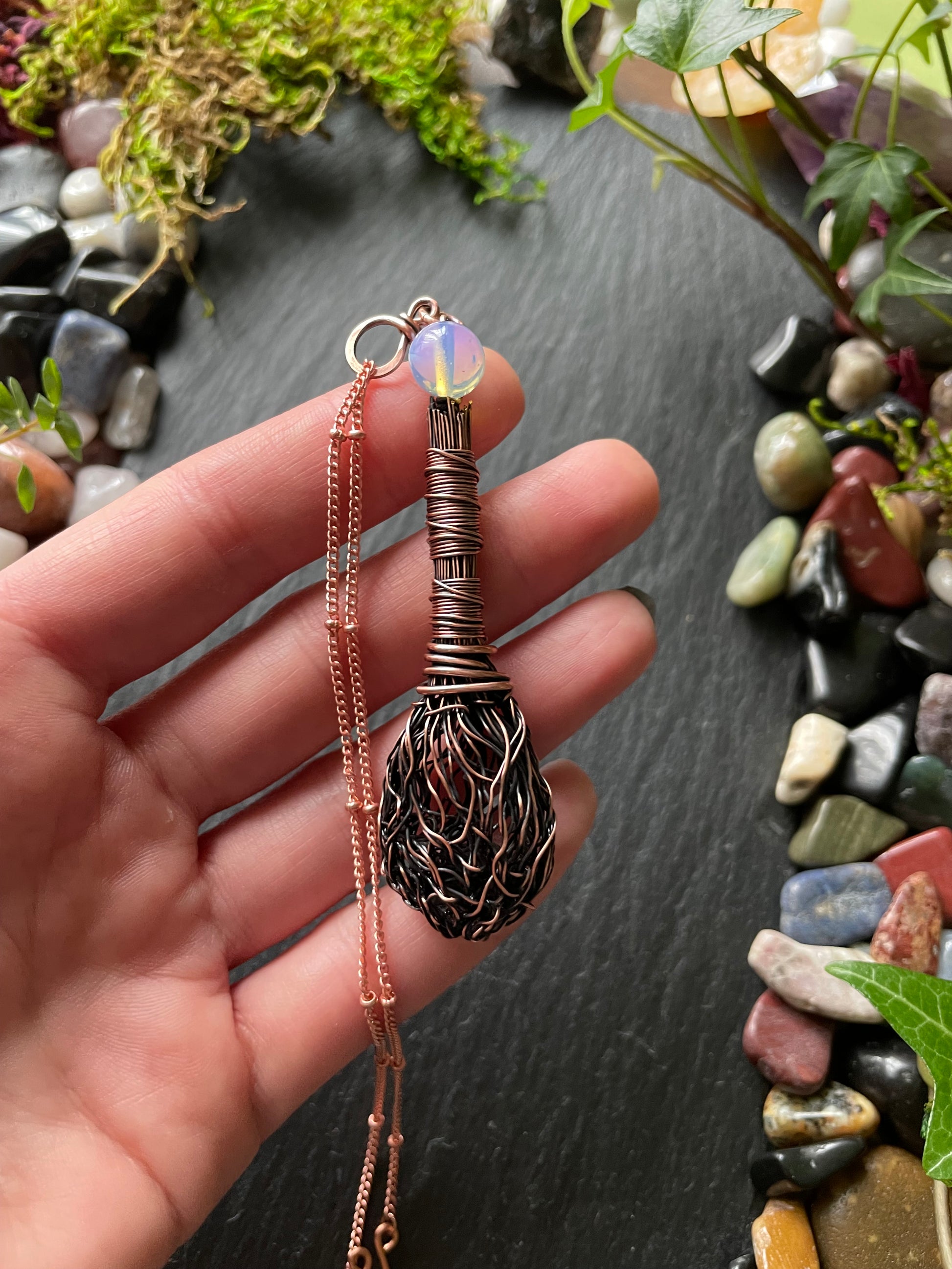 Hand holding a decorative broom-shaped pendant with intricate design on a natural stone background.