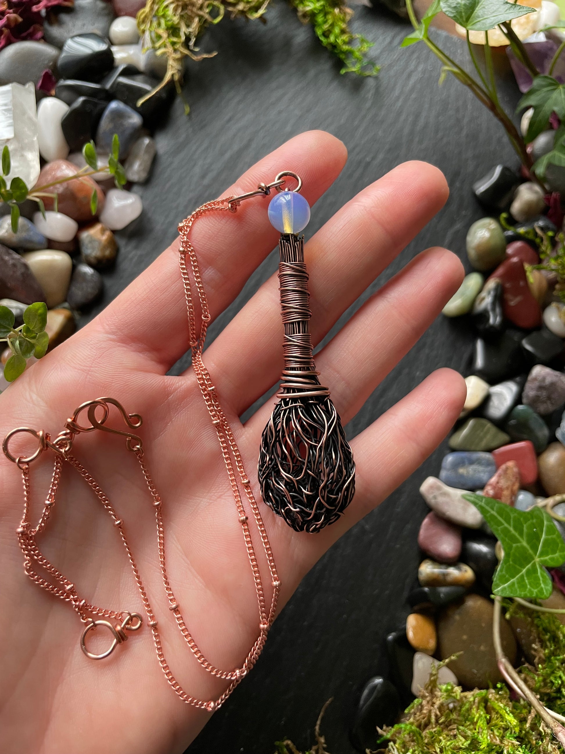 Hand holding a necklace with a decorative pendant against a natural stone and plant background