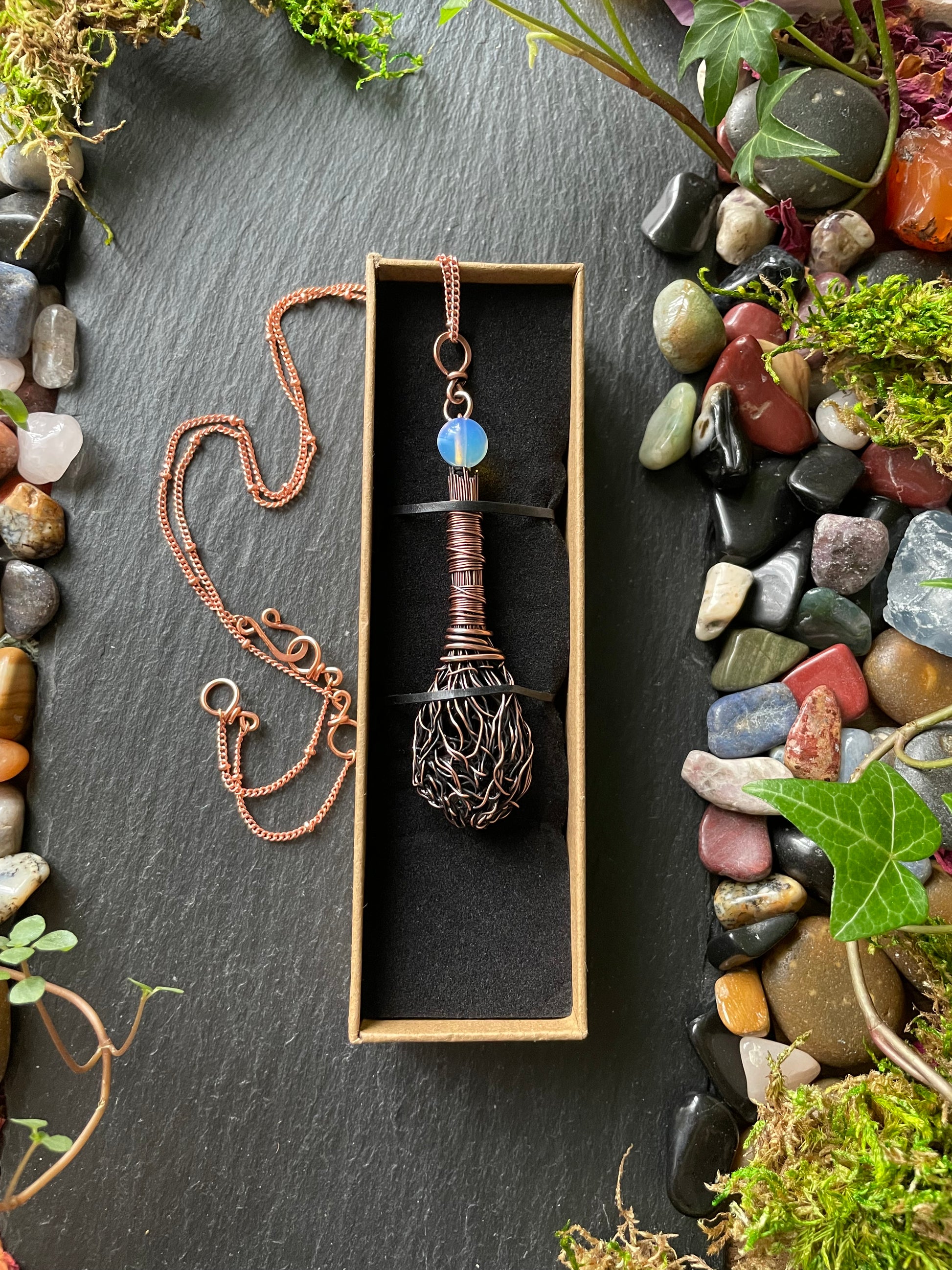 Copper necklace with a blue stone in a box on a dark surface with stones and plants.