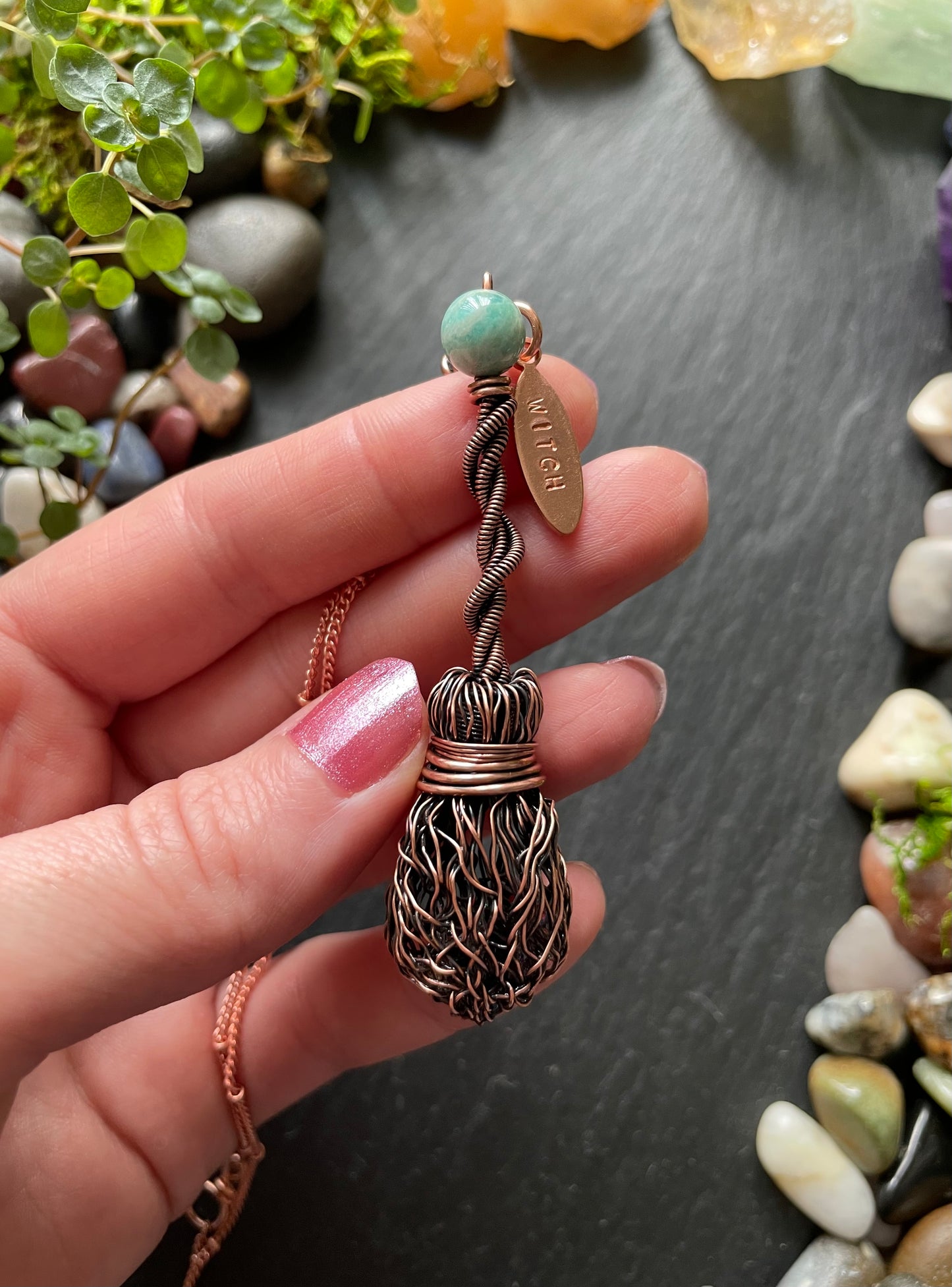 Hand holding a decorative pendant with pebbles and plants in the background