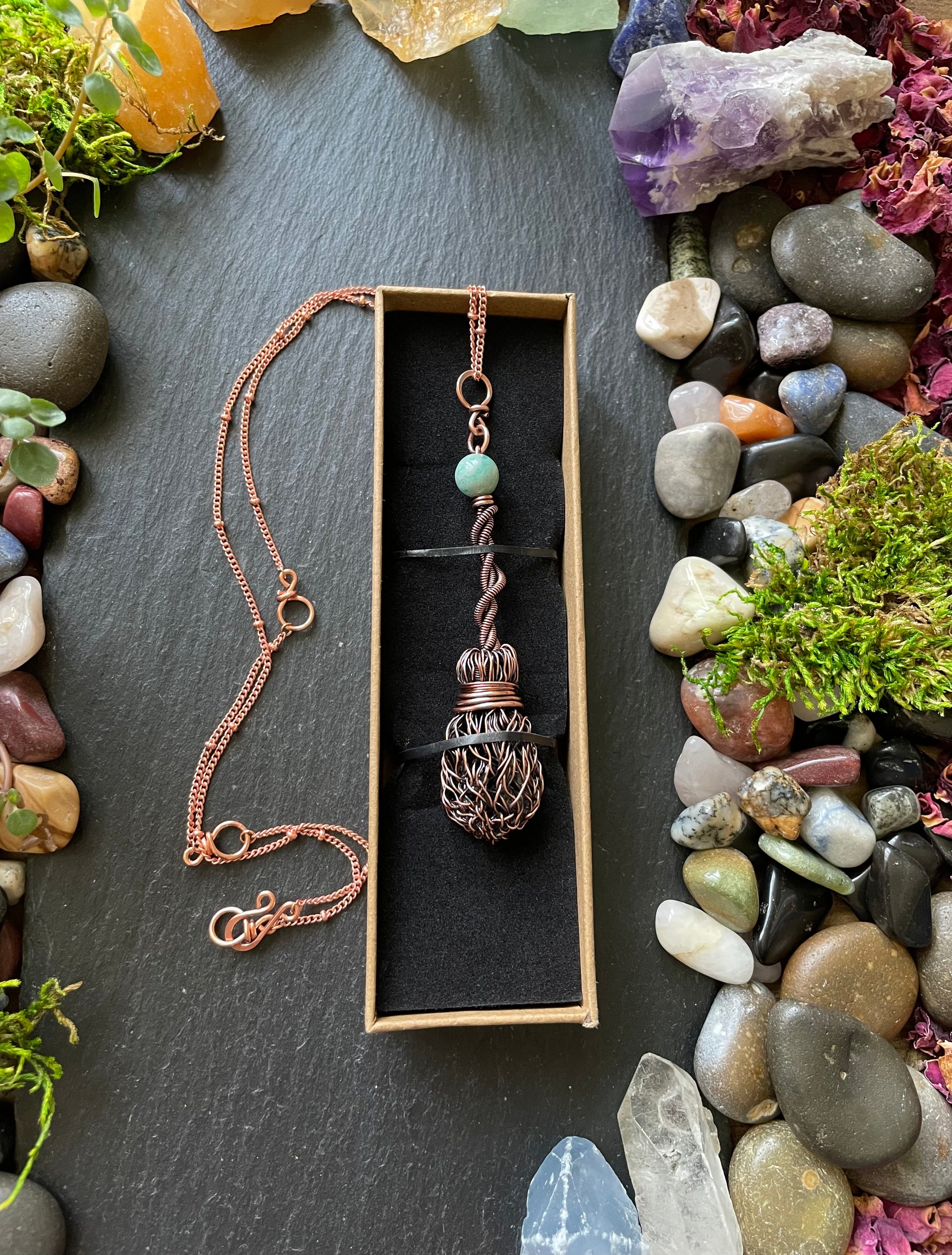 Copper necklace with a pendant in a box, surrounded by stones and plants on a dark surface
