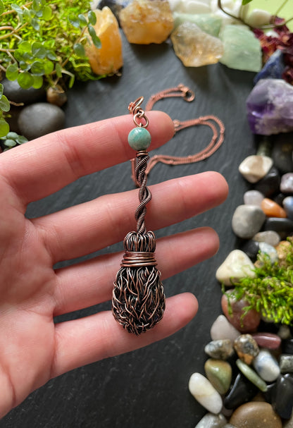 Hand holding a decorative pendant with a green bead against a background of stones and crystals.