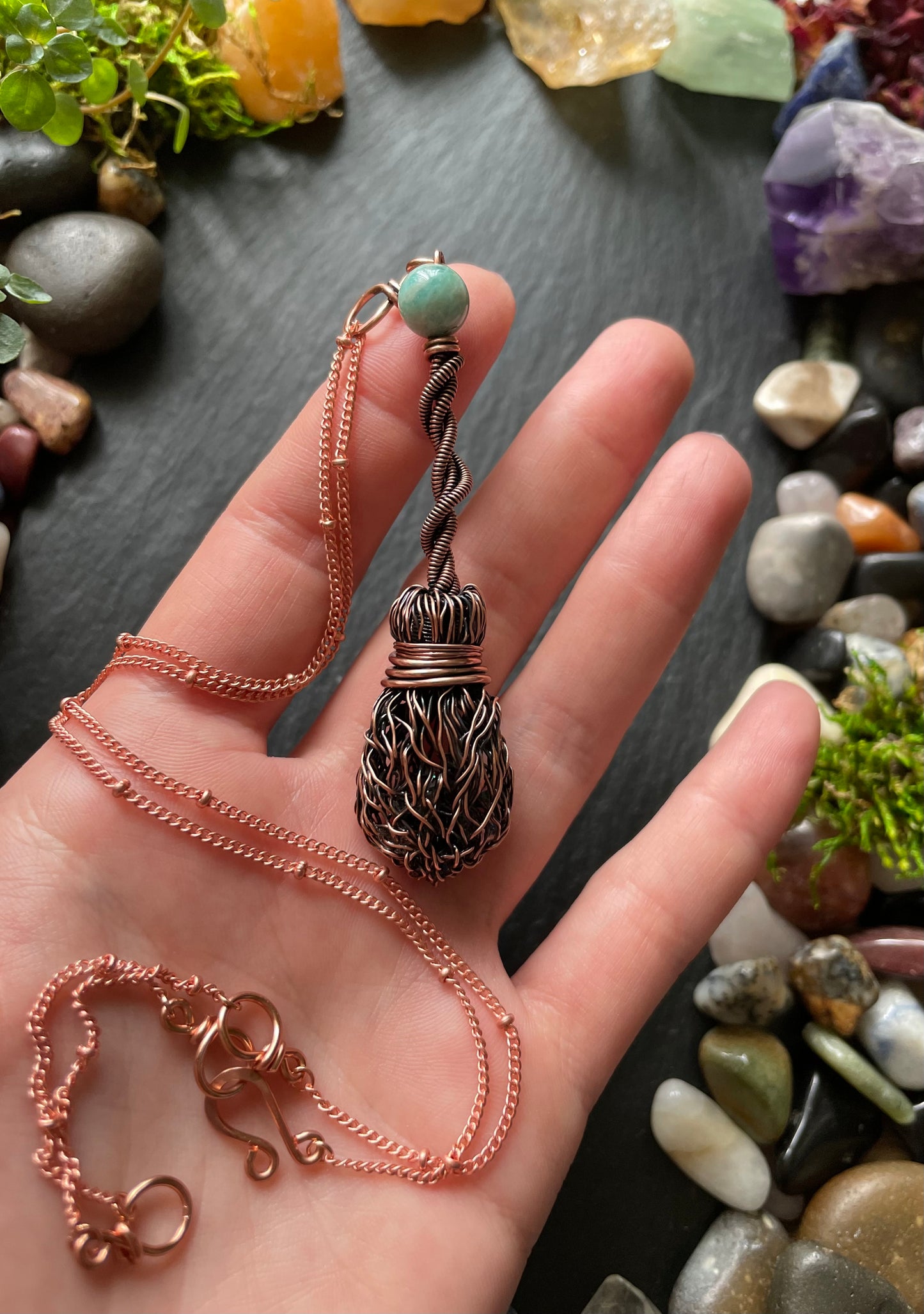 Hand holding a copper necklace with a decorative pendant against a background of stones and crystals.