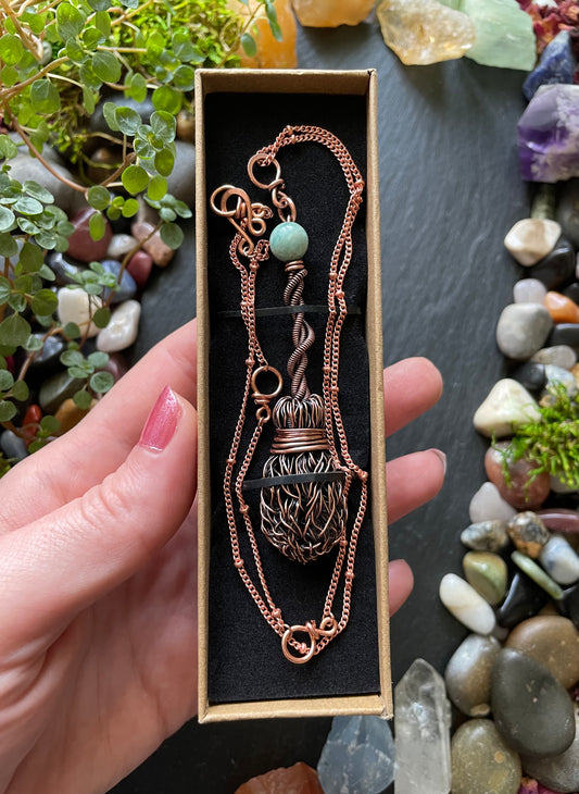 Copper broom necklace with a green gemstone in a box, surrounded by stones and plants.