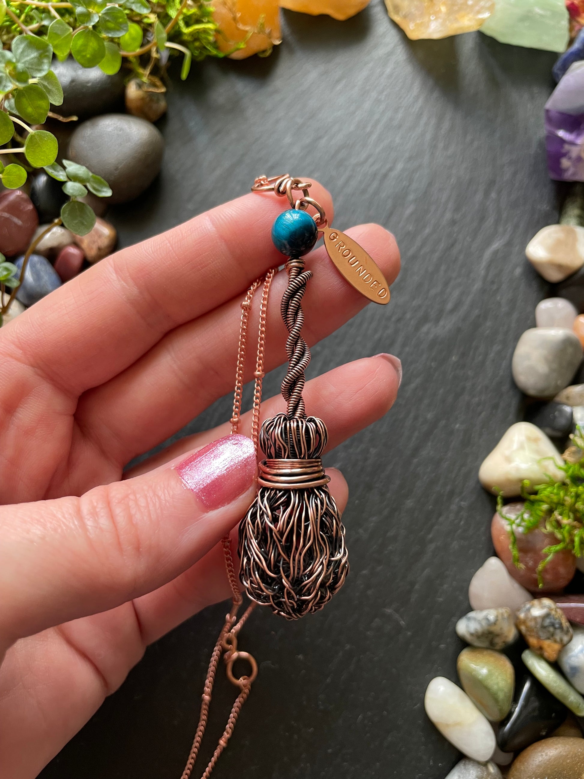 Hand holding a decorative pendant with a textured design and blue bead, set against a natural stone background.