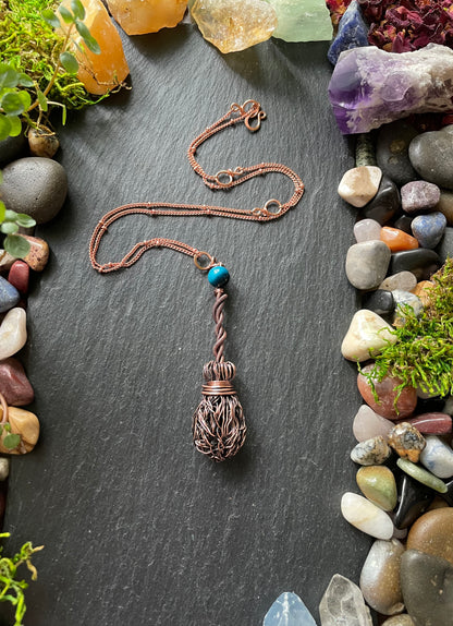 Copper necklace with a pendant on a dark surface surrounded by stones and plants