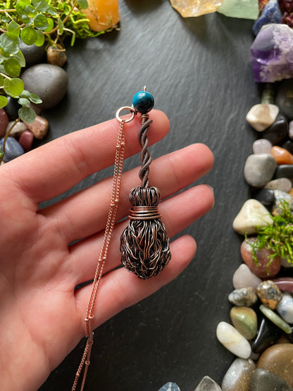 Hand holding a decorative necklace with a copper name tag and bead against a background of stones and plants.