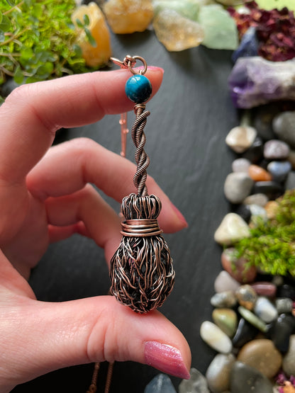 Hand holding a decorative pendant with a textured design against a background of stones and plants