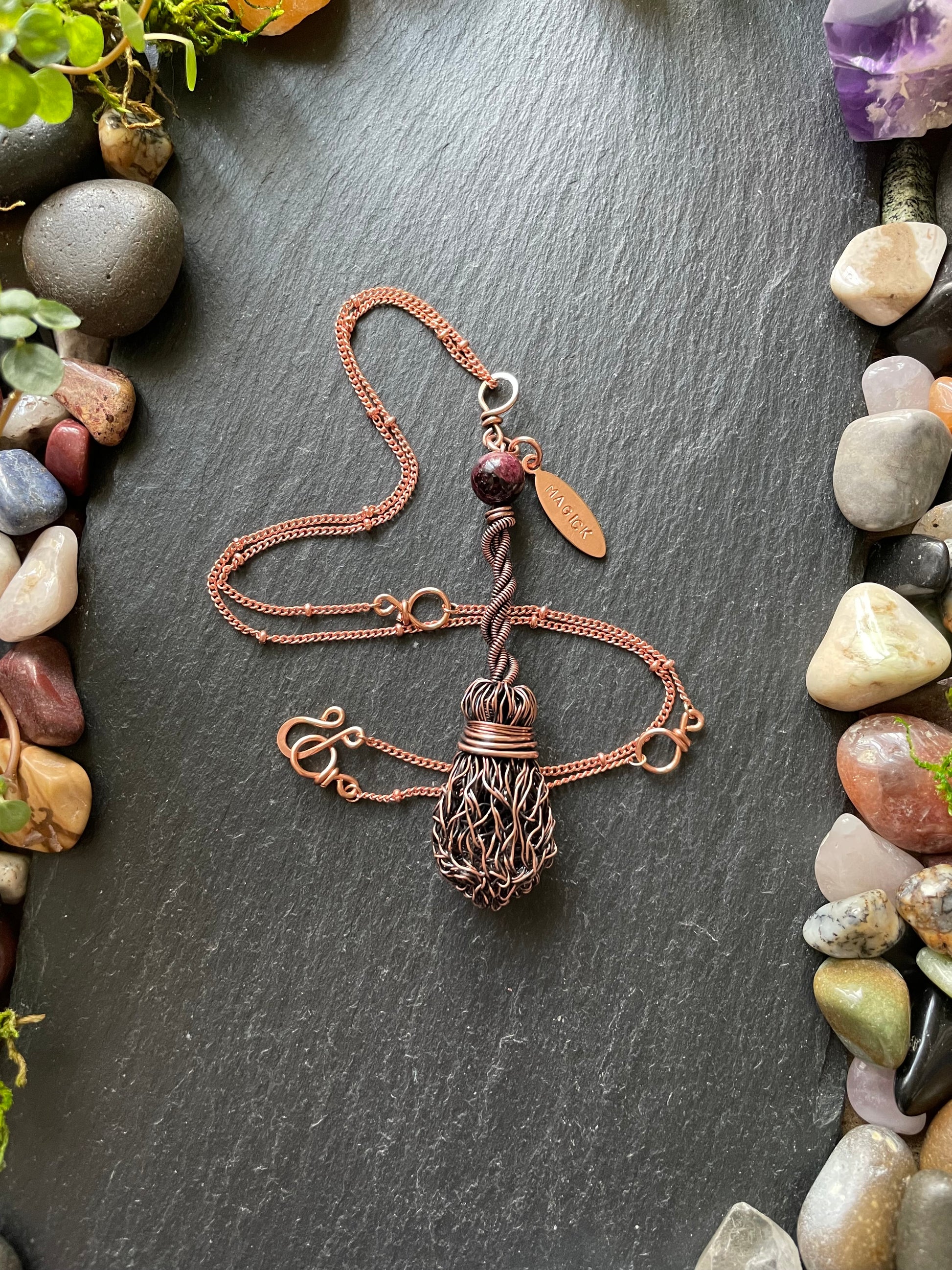 Copper necklace with a magick name tag on a dark stone surface surrounded by pebbles and small plants