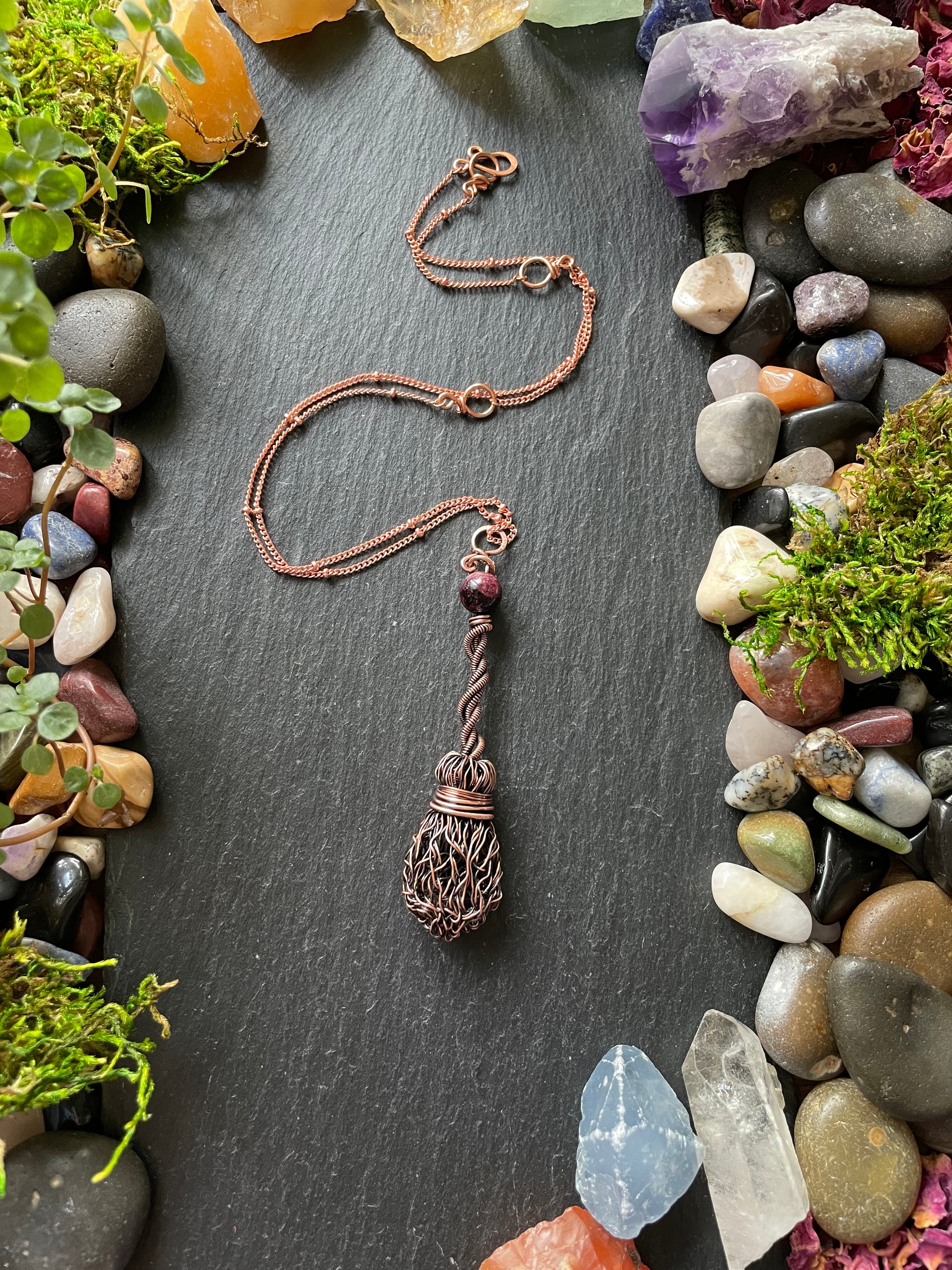 Copper pendant with decorative elements on a dark surface surrounded by stones and plants
