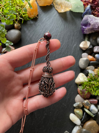 Hand holding a copper necklace with a decorative pendant against a background of stones and plants