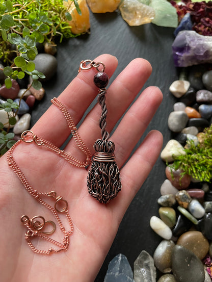 Hand holding a copper necklace with a decorative pendant against a natural stone and plant background