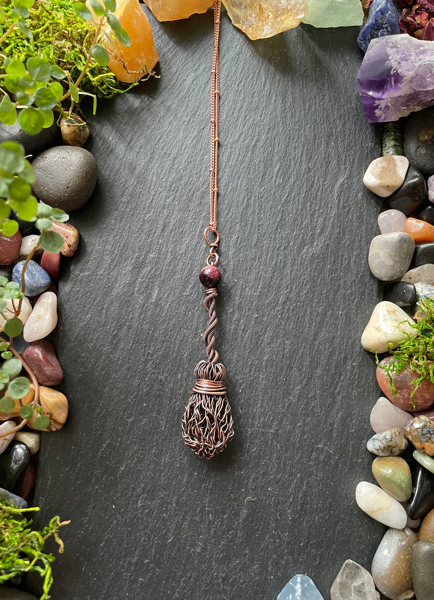 Copper pendant necklace on a dark slate surface with stones and plants
