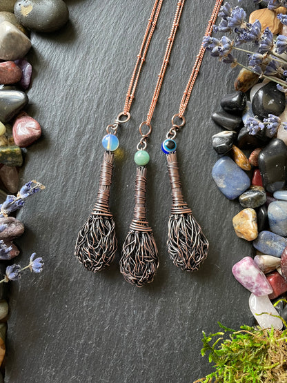 Three miniature copper wire witch broom pendants with opalite, amazonite, and evil eye bead displayed on a stone slate and surrounded by gemstones and moss