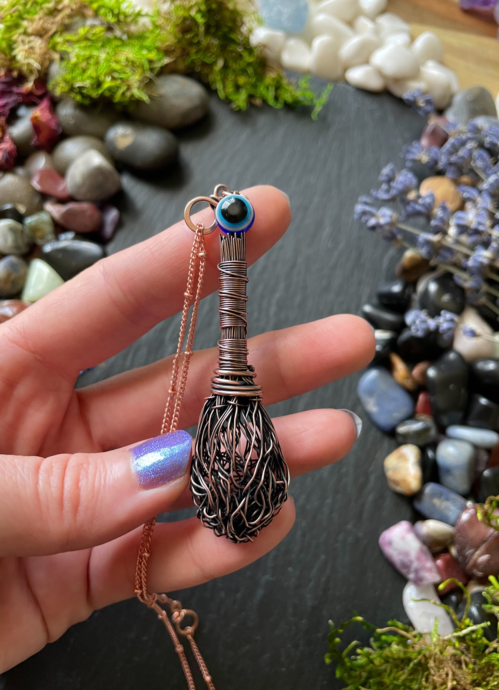 Miniature antiqued copper wire broom pendant with wire wrapped handle and evil eye resin bead, displayed in hand
