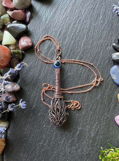 Miniature antiqued copper wire witch broom pendant with resin evil eye bead, attached to a matching copper chain and displayed on a stone slate surrounded by gemstones