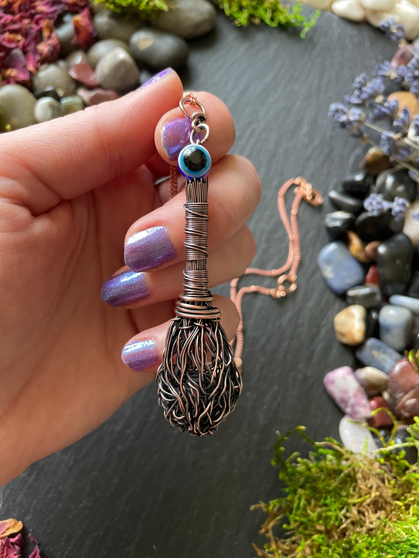 Miniature antiqued copper wire witch's broom pendant with an evil eye bead, displayed in hand