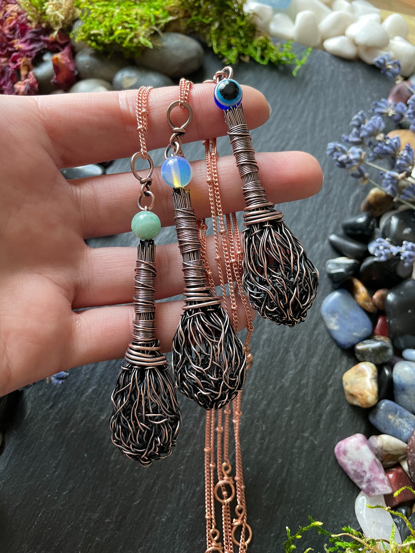 Three miniature antiqued copper wire broom pendants with amazonite, opalite, and evil eye bead, attached to copper chains and displayed in hand