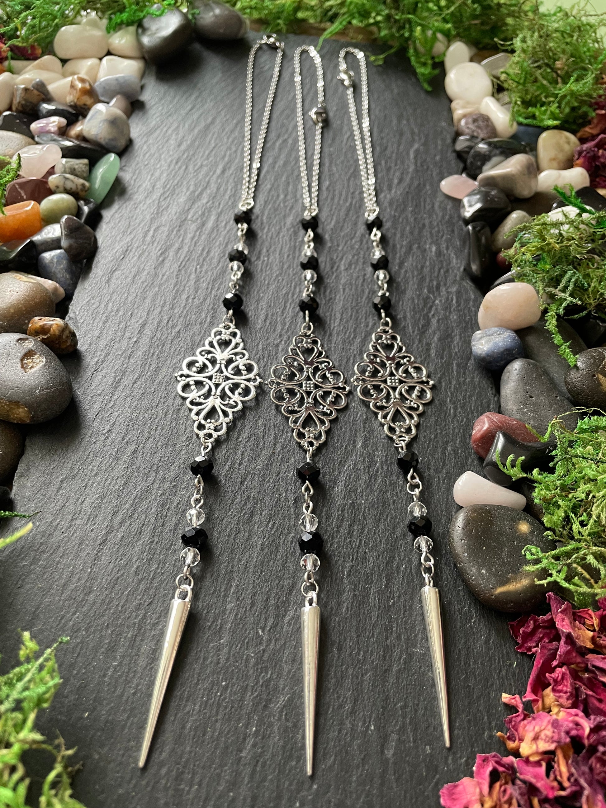 Silver car mirror charms with black beads on a dark stone surface surrounded by stones and plants.